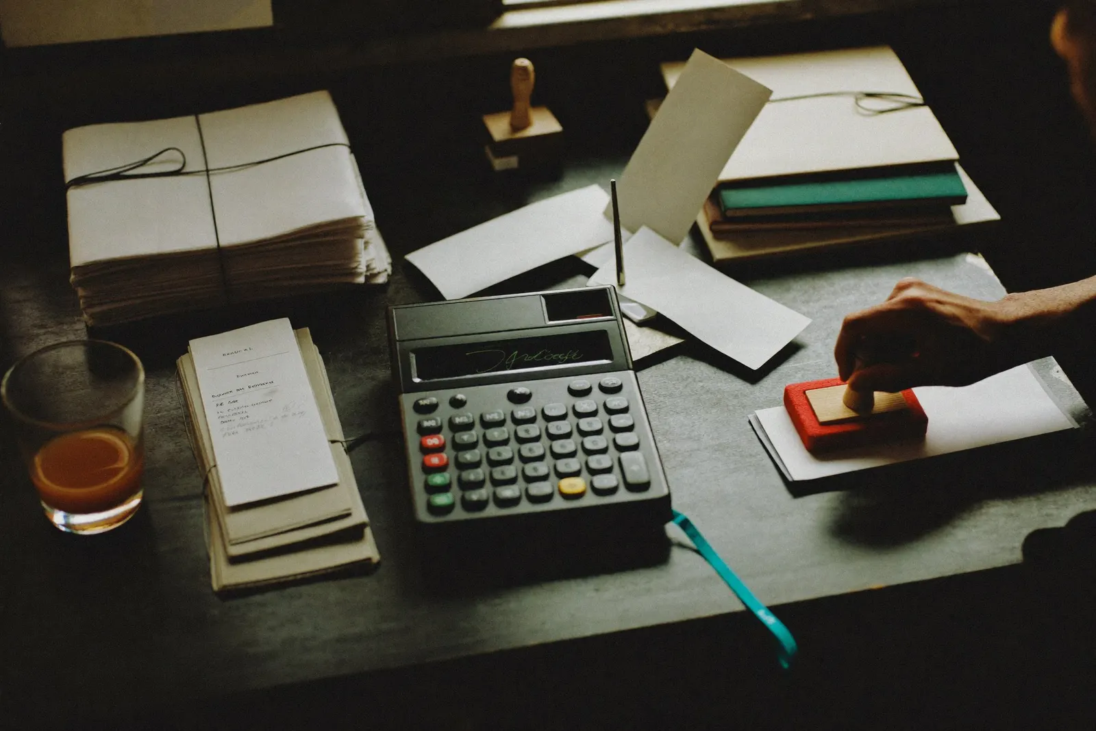 A back-office desk with bundled receipts, a calculator and a stamp in hand