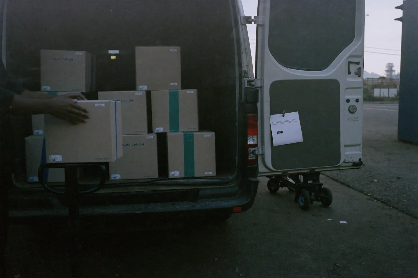 Cardboard boxes stacked in the back of a van with its rear doors open, and a hand placing one more box